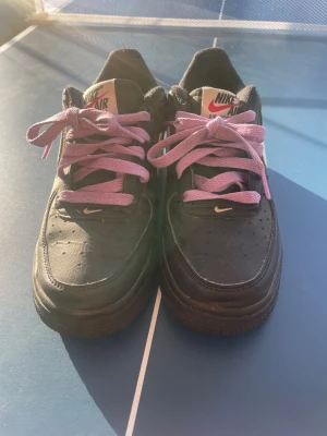 Nike Air svarta sneakers med lila snören - Nike Air sneakers i svart läder med lila skosnören och klassisk Nike-logga på plösen. Medföljer även ett par extra svarta skosnören med röda detaljer. Låg modell med perforerad tåbox för ventilation. Perfekta för en streetwear-look.