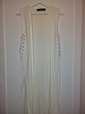 Vit kofta/väst från Zara knit - Elegant vit lång väst från Zara med snygga snörningsdetaljer längs sidorna. Enkel siluett som ger en clean och modern vibe. Inköpt I Spanien och helt slutsåld. Väldigt snygg över jeans.