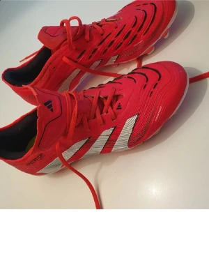 Röda Adidas Predator fotbollsskor - Snygga röda Adidas Predator fotbollsskor med vita detaljer och klassiska tre ränder på sidan. Skorna har snörning, låg profil och är tillverkade i syntetmaterial för bra grepp och kontroll på planen. Perfekta för dig som vill sticka ut på fotbollsplanen.
