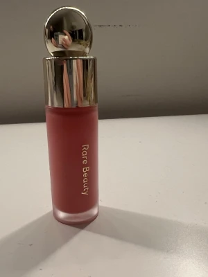 Rare Beauty flytande rouge rosa - Flytande rouge från Rare Beauty i en snygg rosa nyans. Färgen Happy. Kommer i en frostad glasflaska med guldfärgat lock och rund applikator. Perfekt för att ge kinderna en fräsch och naturlig färg. Lätt att blenda och ger ett mjukt resultat. Knappt använd! Mycket kvar med ganska stark pigment!❣️