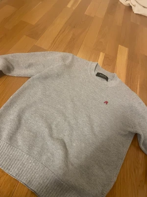 Ljusgrå stickad tröja från Replay - Snygg ljusgrå stickad tröja från Replay med diskret röd broderad 'R' på bröstet. Tröjan har rund halsringning, ribbade muddar och är tillverkad i ett mjukt, strukturerat material som känns skönt mot huden.