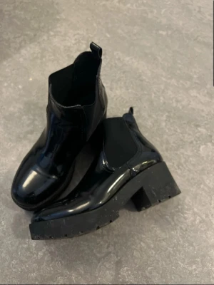 Svarta lackade Chelsea boots Attitude - Säljer ett par svarta Chelsea boots från Attitude med glansig lackad yta och chunky sula. Klassisk resår på sidorna och dragflik bak för enkel påtagning. Perfekta för dig som vill ha en edgy och trendig look.