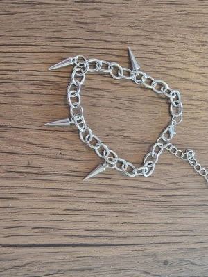 Silvrigt armband med nitar - Säljer ett coolt armband i silverfärgad metall med stora länkar och spetsiga konformade nitar runt om. Armbandet har justerbar kedja och en liten pärldetalj vid låset. 18 cm + 5 cm. Perfekt för dig som gillar edgy accessoarer.