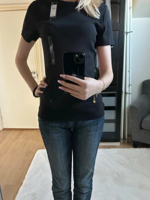 Svart basic t-shirt från Tommy Hilfiger - En stilren svart t-shirt från Tommy Hilfiger med klassisk passform och rund hals. Tillverkad i mjuk bomull som känns skön mot huden. Perfekt till jeans eller shorts för en clean look. Diskret logga på bröstet och korta ärmar. Den ska sitta tajtare men köptes i fel storlek för mig.