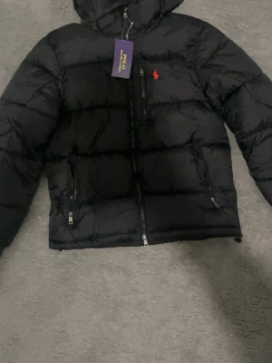 Svart pufferjacka från Polo Ralph Lauren - Säljer en svart pufferjacka från Polo Ralph Lauren med huva och dragkedjor. Jackan har det klassiska röda Polo-logot broderat på bröstet och flera praktiska fickor med dragkedja. Perfekt för kalla dagar och riktigt snygg till streetwear-stilen.