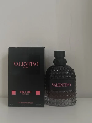 Valentino Uomo Born in Roma Intense - Säljer en lyxig parfym från Valentino, Uomo Born in Roma Intense. Flaskan är svart med en cool, noppad design och rosa detaljer. Parfymen kommer i en elegant svart kartong med rosa text och är perfekt för dig som vill sticka ut med en unik doft.