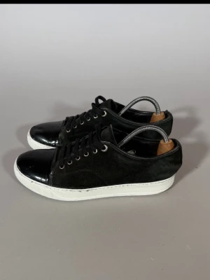 Svarta sneakers med vit sula - Säljer ett par stilrena svarta sneakers med vit sula. Skorna har en kombination av mocka och glansigt material på tån och hälen, samt svarta skosnören och metallöljetter. Perfekta för dig som gillar en clean och klassisk look.