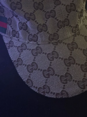 Gucci keps med GG-mönster - Säljer en beige och brun keps från Gucci med det klassiska GG-monogrammet över hela kepsen. På sidan finns en grön och röd rand som signaturdetalj. Kepsen är tillverkad i canvasmaterial och har böjd skärm. Perfekt för dig som gillar exklusiva accessoarer.