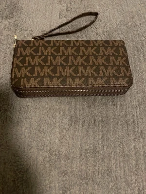 Brun MK plånbok med handledsrem - Snygg brun plånbok från Michael Kors med klassiskt MK-mönster över hela ytan. Plånboken har en praktisk handledsrem och dragkedja runt om. Tillverkad i tåligt konstläder och har en rektangulär form som rymmer kort, kontanter och mobil.