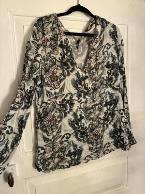 Mönstrad blus med v-ringning - Snygg blus från Odd Molly med v-ringning och långa ärmar. Blusen har ett unikt, färgstarkt mönster i svart, grått och röda detaljer på ljus botten. Tillverkad i ett tunt, lätt tyg som ger en luftig känsla. Perfekt för dig som gillar bohemisk stil. Storlek 3 som motsvarar ca L/42 i EU