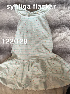 Randig volangklänning med prickar - Söt klänning med ljusblå och vita ränder samt små färgglada prickar. Klänningen har volang nertill och är ärmlös med rund halsringning. Perfekt för varma dagar och ger en lekfull look.🌸Små mindre synliga fläckar. Storlek 122/128  