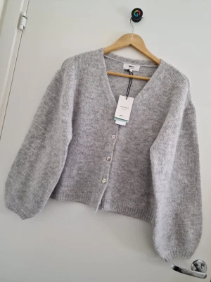 Ljusgrå kofta från Gina Tricot S - Mysig ljusgrå kofta från Selected Femme med v-ringning och tre knappar framtill. Koftan har en avslappnad passform, ballongärmar och är stickad i ett mjukt material som ger en fluffig känsla. Perfekt för lager på lager och enkel att styla. Storlek S.