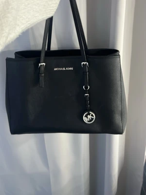 Svart Michael Kors handväska - Stilren svart handväska från Michael Kors i slätt konstläder med silverfärgade detaljer. Väskan är i bra kvalitet men är lite sliten på banden. 💕