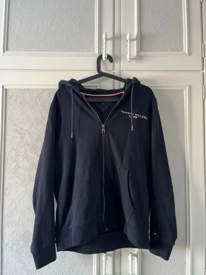 Svart hoodie från Tommy Hilfiger - Snygg svart hoodie från Tommy Hilfiger med dragkedja framtill, huva med dragsko och två stora fickor. Diskret logga på bröstet och klassisk design. Perfekt för en avslappnad och stilren look.
