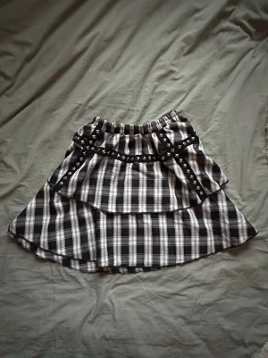 Svartvitrutig kjol med nitar - Säljer en svartvitrutig kjol med lager på lager-design och elastisk midja. Kjolen har ett band med nitar i för en edgy look. Perfekt för dig som gillar alternativ stil och vill sticka ut. Står ingen storlek på, med stretchig och passar många! Själv bär jag storlek xs, sp skulle säga att den passar s bäst.⭐️