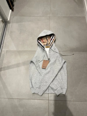 Grå zip hoodie från Burberry 100 procent cotton - Snygg grå zip hoodie från Burberry med klassiskt rutigt foder i huvan. Hoodien har dragkedja framtill, ribbade muddar och justerbar dragsko i huvan. Perfekt för dig som vill ha en stilren och exklusiv look med ikoniska Burberry-detaljer.