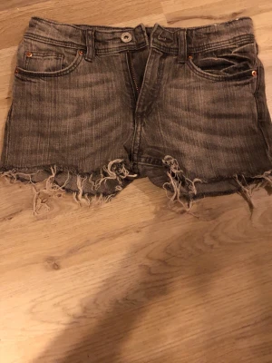 Grå jeansshorts med fransig kant H&M - Säljer ett par grå jeansshorts från H&M med fransig, rå kant och klassisk femficksdesign. Shortsen har midwaist och knappgylf. Perfekta för en avslappnad och trendig look.
