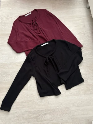 Två knytkoftor i vinrött och som strl S - Två trendiga koftor med knytning framtill, en i vinrött och en i svart. Båda har rund halsringning och långa ärmar. Perfekta att slänga över en topp för en chill look. Materialet ser ut att vara mjukt och stretchigt.