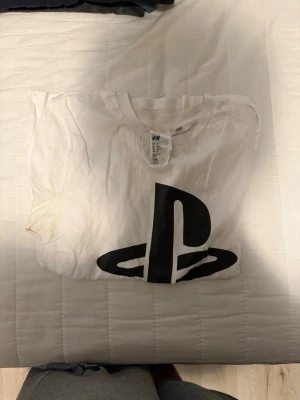 Vit PlayStation t-shirt från H&M - Säljer en vit t-shirt från H&M med stor svart PlayStation-logga på framsidan. Klassisk rund hals och korta ärmar. Perfekt för dig som gillar gaming och vill ha en enkel men snygg look.
