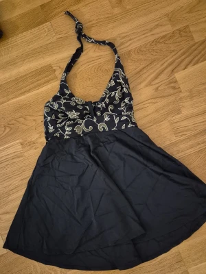 Svart baddräkt med halterneck - Snygg svart baddräkt med halterneck och kjol. Överdelen har ett blommigt mönster i vitt och knytning i nacken. Kjoldelen är enfärgad svart och ger en retro känsla. Perfekt för strandhäng eller poolparty.