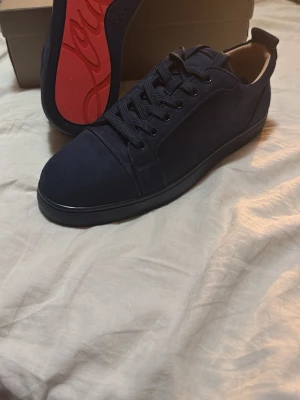 Louboutins mörkblå mocha  - Snygga marinblå sneakers från Christian Louboutin med klassisk låg profil och signaturröd sula. Skorna är gjorda i mocka och har matchande marinblå skosnören samt broderad logga på hälen. Perfekta för dig som vill ha en stilren och exklusiv look. Helt nya