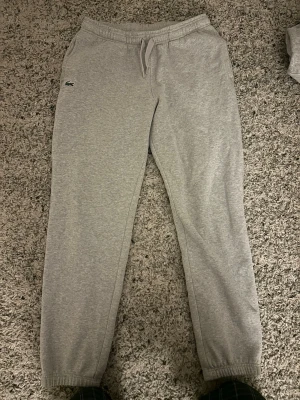 Grå mjukisbyxor från Lacoste - Säljer ett par grå mjukisbyxor från Lacoste med klassisk krokodillogo på vänster sida. Byxorna har dragsko i midjan, sidofickor och muddar vid bensluten. Perfekta för chill dagar eller när du vill ha en clean och enkel look.