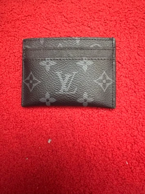 Louis Vuitton svart kortfodral - Snyggt svart kortfodral från Louis Vuitton med klassiskt monogrammönster i grått. Tillverkat i läder och har flera fack för kort. Perfekt för dig som vill ha en stilren och lyxig accessoar i fickan.