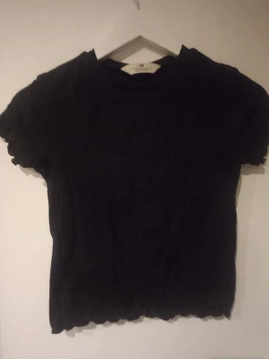 Svart ribbad topp från H&M - En svart ribbad topp från H&M med korta ärmar och vågiga kanter vid ärmslut och nederkant. Toppen har en enkel, tight passform och är perfekt för att styla med jeans eller kjol. Materialet är mjukt och stretchigt för extra komfort.