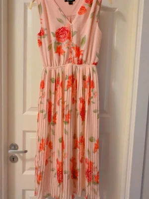 Blommig långklänning i rosa toner - Söt långklänning med blommigt mönster i rosa, rött och grönt. Klänningen har v-ringning, knappar framtill och är ärmlös. Perfekt för varma dagar och har ett lätt, luftigt tyg som ger en romantisk känsla.