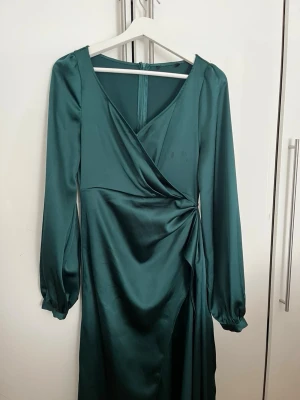 Mörkgrön omlottklänning i satin - Snygg mörkgrön omlottklänning i glansigt satintyg med långa ärmar och v-ringning. Klänningen har en draperad detalj i midjan och en slits. Perfekt för dig som vill ha en stilren och trendig look.