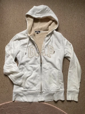 Vintage y2k pälsfodrad teddy zip hoodie - Vit vintage hoodie från GAP med broderad logga framtill. Mjuk teddyfoder på insidan och huva med snören. Mycket mysig och gosig isolerad! Liknar päls hoodies, men detta är lättare att sköta om💕Dragkedja hela vägen och två fickor framtill. Perfekt för kyliga dagar och riktigt cozy vibes!😻Väldigt populär och sällsynt💋Så snygg y2k 2000s zip hoodie!! Storlek S, passar XS också. Använd några gånger, inga fel! Bara lite lite fläckig vid kragen framtill🩷Hör av dig vid frågor, pris kan diskuteras❤️