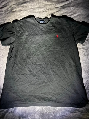 Svart t-shirt från Ralph Lauren - Klassisk svart t-shirt från Ralph Lauren med rund hals och korta ärmar. Liten röd broderad logga på bröstet ger en stilren touch. Tillverkad i mjuk bomull för skön känsla hela dagen.