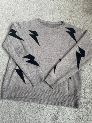 Grå stickad tröja med blixtmotiv - Säljer en grå stickad tröja med svarta blixtar som detaljer. Tröjan har rund halsringning, långa ärmar och ribbade muddar vid ärmslut och nederkant. Perfekt för dig som gillar en cool och avslappnad stil. Den har en litet fläck på högra armbågen som inte är något man tänker på, visas på sista bilden. 