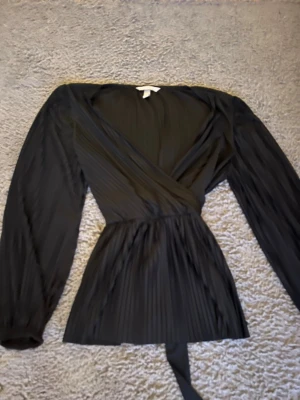 Svart omlottblus med plissering - Snygg svart omlottblus med plisserat tyg och knytband i midjan. Blusen har långa ärmar och v-ringad halsringning som ger en elegant look. Perfekt att styla med jeans eller kjol för en trendig outfit.