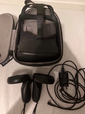 Oculus Quest VR-headset med tillbehör - Säljer ett Oculus Quest VR-headset i svart med tillhörande handkontroller, laddkabel och en praktisk grå förvaringsväska. Headsetet har justerbart band och bekväm vaddering för längre spelsessioner. Perfekt för dig som vill uppleva VR-spel för första gången. BeatSaber är riktigt roligt men det finns många fler spel att upptäcka.