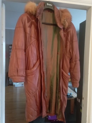 Brun lång skinnjacka med luva - Lång brun skinnjacka från Emary med stor luva och fluffig fuskpäls runt huvan. Jackan har två stora fickor framtill, dekorativa sömmar och en unik detalj på bröstet. Perfekt för dig som vill ha en statementjacka med retrovibbar. Tyvärr är fodret trasigt men går att sy ihop. 2 vita fläckar på framsidan ovanför ena fickan.