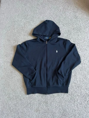 Mörkblå hoodie från Polo Ralph Lauren - Snygg mörkblå hoodie från Polo Ralph Lauren med dragkedja framtill och klassisk vit logga på bröstet. Hoodien har två fickor fram och en skön huva. Tillverkad i mjukt bomullsmaterial som passar perfekt till chill vardagsstil.