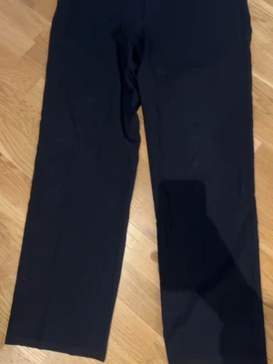 Armani kostymbyxor - Säljer ett par svarta Armani Collezioni kostymbyxor med rak passform och klassisk look. Byxorna har enkel design och är tillverkade i ett slätt bomull som ger en stilren känsla. Perfekta för dig som gillar minimalistisk stil.