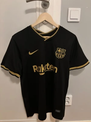 Svart FC Barcelona matchtröja Nike - Snygg svart FC Barcelona matchtröja från Nike med guldfärgade detaljer och logga. Tröjan har V-ringning med guldkant, korta ärmar och Rakuten-tryck på bröstet. Perfekt för dig som älskar fotboll och vill ha en stilren supportertröja.