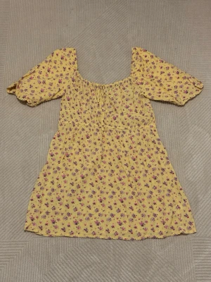 Gul blommig kortklänning med puffärm - Söt gul kortklänning med små blommor i rosa och lila. Klänningen har puffiga korta ärmar, smock i midjan och en söt knytning framtill. Perfekt för varma dagar och ger en romantisk vibe.
