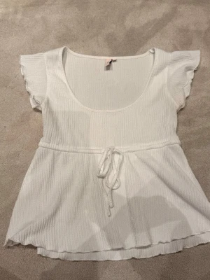 Vit topp med knyt och volangärm - Supersöt vit topp med korta volangärmar och rundad urringning. Toppen har ett knytband i midjan som ger en fin siluett och lätt rynkad nederkant. Materialet är ribbat och känns mjukt mot huden