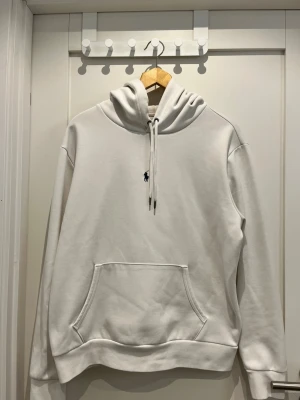 Vit hoodie från Polo Ralph Lauren - Snygg vit hoodie från Polo Ralph Lauren med klassisk huva. Har en stor känguruficka framtill och den ikoniska mörkblå loggan broderad på bröstet. Tillverkad i mjuk bomullsblandning som känns riktigt skön mot huden. Perfekt för en clean och stilren look. Nypris 2100kr