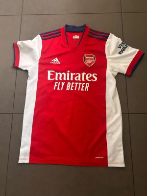 Arsenal matchtröja Adidas röd/vit - Säljer en officiell Arsenal matchtröja från Adidas i rött och vitt med klubbmärke på bröstet och 'Emirates Fly Better' tryck på framsidan. Tröjan har blå detaljer vid kragen och axlarna samt ventilationsmaterial för extra komfort.