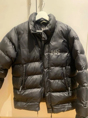 Svart pufferjacka från Helly Hansen - Säljer en svart pufferjacka från Helly Hansen med hög krage och två sidofickor. Jackan har en diskret HH-logga på bröstet och är tillverkad i ett glansigt material som ger en modern look. Perfekt för kalla vinterdagar och riktigt skön att bära.