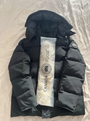 Svart dunjacka från Canada Goose - Säljer en svart dunjacka från Canada Goose med avtagbar huva och patch-logga på ärmen. Jackan har en klassisk puffer-modell och medföljer en lös pälskrage. Perfekt för kalla vinterdagar och riktigt varm.
