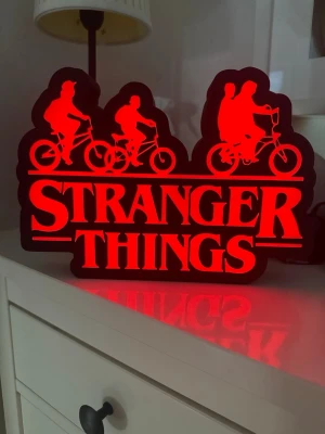 Stranger Things LED-lampa dekoration - Säljer en snygg Stranger Things LED-lampa i svart och rött. Lampan har formen av loggan från serien med siluetter sopplande cyklar ovanför texten. Perfekt som cool inredningsdetalj för fans av serien. Tillverkad i plast och lyser upp med starkt rött sken.