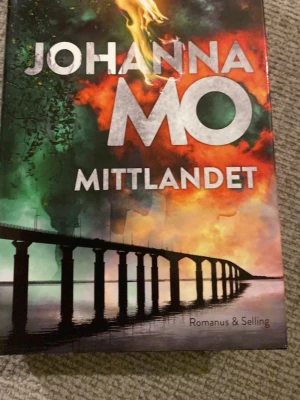 Mittlandet av Johanna Mo - En spännande skönlitterär bok med färgstarkt omslag i grönt, orange och svart. Titeln 'Mittlandet' och författaren Johanna Mo syns tydligt. Omslaget har en dramatisk bro mot en färgstark himmel. Perfekt för dig som gillar deckare och spänning.