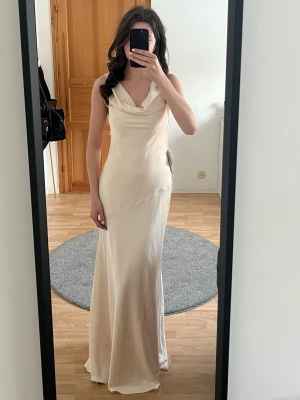 Lång champagnefärgad klänning - Elegant långklänning i champagnefärg med öppen rygg med knyt. Klänningen har en figurnära passform och är tillverkad i ett glansigt, mjukt material som ger en lyxig känsla. Jag är 174cm och bilden är tagen utan skor. Aldrig använd, båda orginalförpackning och prislapp är kvar. 