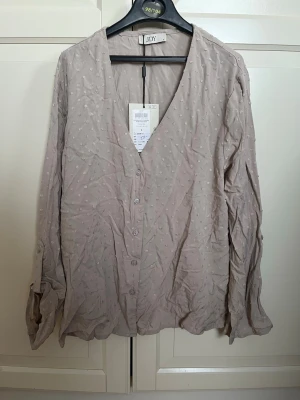 Beige prickig blus från JDY - Snygg beige blus från JDY med diskreta små prickar i tyget. Blusen har v-ringning, knäppning framtill och långa ärmar. Materialet ser lätt och luftigt ut, perfekt för en avslappnad men ändå stilren look.