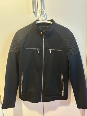 Zara Svart bikerinspirerad jacka med dragkedjor - Säljer en svart jacka med bikerstil, quiltade detaljer på axlar och ärmar samt fyra silvriga dragkedjefickor framtill. Jackan har ståkrage med knapp och är gjord i ett grovt tyg som ger en cool look. Perfekt för dig som gillar streetwear och vill sticka ut.